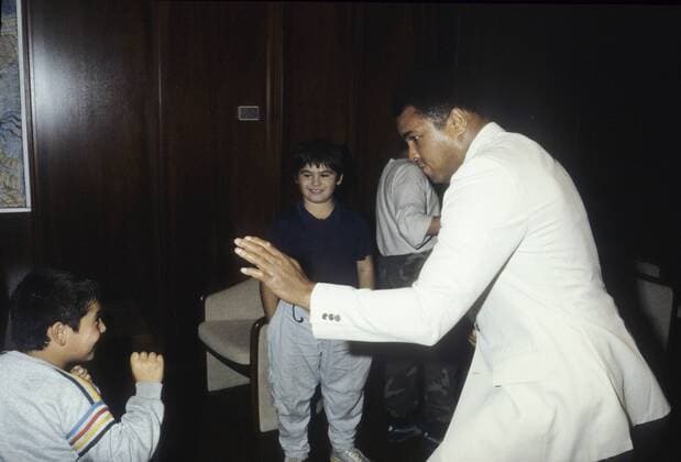 Muhammad Ali (re., Ex-Boxprofi) 09 84 wot Mann ehemaliger Boxer Box-Profi Profi-Sport Profisport Boxen Weltmeister Boxweltmeister dunkelhäutig dunkelhaarig kurzhaarig seriös Jackett Anzug Hemd weiss Krawatte stehend Saal Halle halb quer spielerisch boxend mit einem Kind Fan Cassius Clay