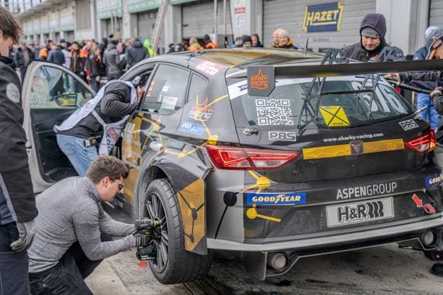 In the pit lane of the Nürburgring, a mechanic from sharky racing com works on the rear wheel of the black and gold Cupra racing car with a pneumatic screwdriver The vehicle bears the starting number 821 and is decorated with sponsors such as Goodyear, H R, Aspen Group and PBS In the background, spectators and team members crowd along the pit wall while other crew members work on the drivers door sharky racing com mechanic changing tires in the pit lane at the Nürburgring Copyright: xspeedshotx