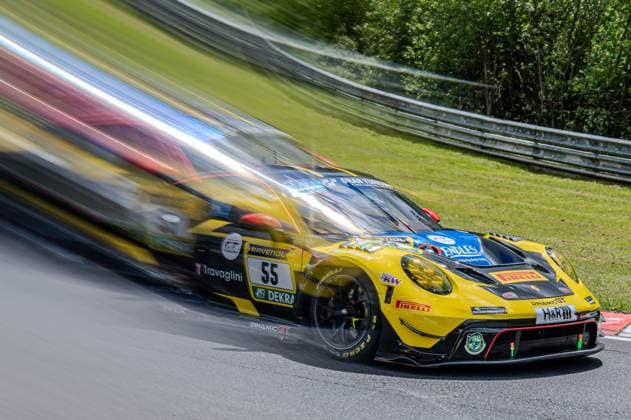The yellow and black Porsche 911 GT3 R from Dinamic GT with starting number 55 Alessandro Ghiretti Joel Sturm Loek Hartog Michele Beretta Dinamic GT races through a bend on the Nordschleife at the Nürburgring with impressive momentum The panning shot creates a strong motion blur effect, which emphasizes the enormous speed of the vehicle Sponsors such as RAVENOL, DEKRA, Travaglini, Pirelli, KW, H R and Endles are clearly visible on the bodywork A crash barrier and green vegetation of the Eifel can be seen in the background Dinamic GT Porsche 55 at full speed on the Nordschleife Copyright: xspeedshotx