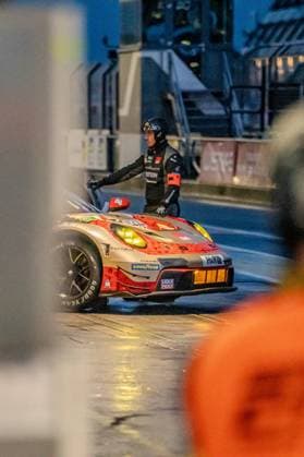 In the Nürburgring pit lane at night, a mechanic in black racing overalls and helmet stands on the Lukas Ertl Matthias Benndorf Maximilian Johannes Ertl Stefan Ertl Reiter Engineering - a Porsche 911 GT3 R from Reiter Engineering with the starting number 91 The vehicle has a striking gold and red livery with sponsors such as LIQUI MOLY, Goodyear, Fanatec and H R The yellow fog lights shine in the rainy environment The mechanic holds on to the open roof, possibly during a vehicle change or an inspection in the pits Porsche 911 GT3 91 by Reiter Engineering in the pit lane at night Copyright: xspeedshotx
