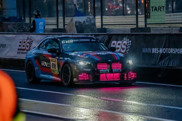 The BMW M2 with the starting number 898 from Walkenhorst Motorsport Bennet Ehrl Maxim Felix Dacher Takayuki Kinoshita Tazio Ottis Walkenhorst Motorsport drives on wet asphalt at the Nürburgring at night The car has a dark paint job with characteristic BMW Motorsport stripes in red and blue as well as striking pink magenta LED lighting on the radiator grille and front apron The EHR Gran Turismo lettering can be seen on the windshield, and sponsors such as H R and DEKRA BBS Motorsport advertising banners and the START sign are visible in the background - the vehicle is apparently on the starting grid or in the pit lane BMW M2 898 from Walkenhorst Motorsport at night at the Nürburgring Copyright: xspeedshotx