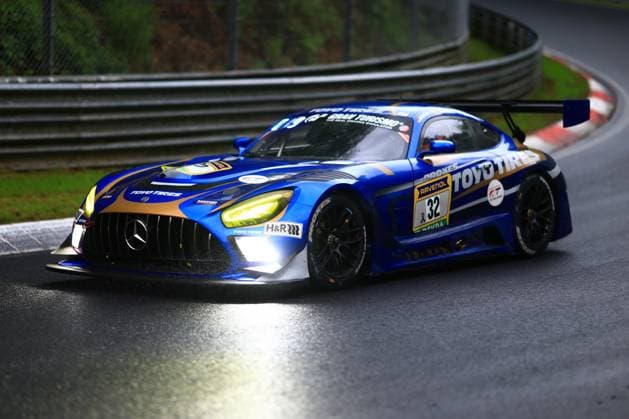 The Mercedes AMG GT3 with starting number 32 of the Toyo Tires with Ring Racing team drives through a wet corner on the Nordschleife of the Nürburgring The vehicle Andreas Gülden Tim Sandtler Yuichi Nakayama Toyo Tires with Ring Racing wears a striking blue and gold livery with the sponsor logos of Toyo Tires, RAVENOL, Gran Turismo and H R The switched-on LED headlights reflect on the damp asphalt while the car is steered precisely through the exit of the corner Mercedes AMG GT3 32 by Toyo Tires with Ring Racing on the Nordschleife Copyright: xspeedshotx