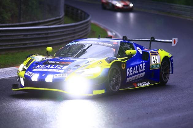 The Ferrari GT3 with the starting number 45 from REALIZE KONDO RACING with Rinaldi drives through a corner on the Nürburgring Nordschleife in damp, dusky weather The car David Perel Dennis Marschall Thierry Vermeulen Thomas Neubauer REALIZE KONDO RACING with Rinaldi presents an eye-catching blue and yellow livery with neon green accents and the sponsors REALIZE GROUP, Rinaldi Racing, RAVENOL, YOKOHAMA, H R and DEKRA The headlights shine brightly on the wet asphalt In the background you can see another car Ferrari 45 from REALIZE KONDO RACING with Rinaldi on the Nordschleife Copyright: xspeedshotx