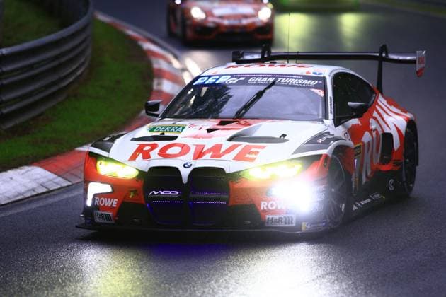 The BMW M4 GT3 Augusto Farfus Jordan Pepper Kelvin van der Linde Raffaele Marciello ROWE RACING from ROWE RACING in the striking red and white livery with large ROWE lettering drives through the Nordschleife of the Nürburgring on a wet track and at dusk The vehicle is presented with its headlights switched on, visible sponsors such as H R and MCG as well as Gran Turismo branding on the windshield Another vehicle can be seen in the background following the BMW M4 GT3 1 from ROWE RACING through the curved section of the track on the Nordschleife Copyright: xspeedshotx