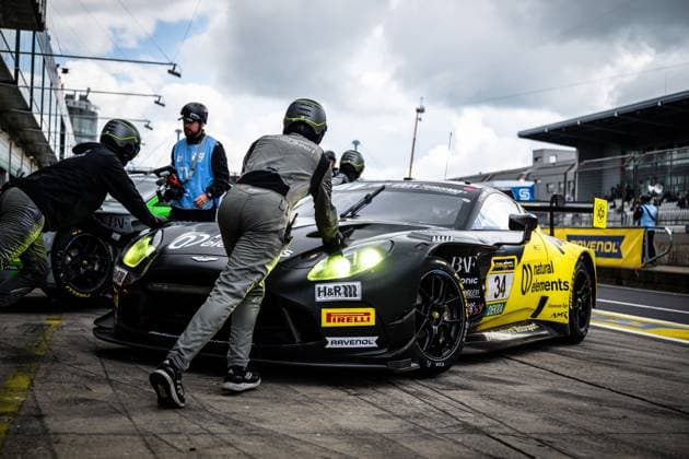 The Aston Martin Vantage GT3 with starting number 34 of the natural elements by Walkenhorst Motorsport team is parked in the pit lane at the Nürburgring Several mechanics in dark overalls and helmets are working intently on the car while a marshal in a blue vest observes the action The black and yellow car bears sponsors such as RAVENOL, Pirelli and H R and is clearly recognizable with its bright headlights Another car can be seen in the pit lane in the background Christian Krognes Mattia Drudi Nicki Thiim natural elements by Walkenhorst Motorsport Aston Martin 34 from natural elements by Walkenhorst Motorsport in the pits Copyright: xMichaelxGelhardtx