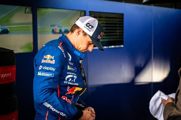 In the pit at the Nürburgring, a driver in a blue racing overall with Red Bull , Viaplay , Michelin and Intercontinental GT Challenge logos can be seen The driver looks down intently and adjusts his glove In the background, monitors with live racing images and a blue pit wall can be seen The drivers AlphaTauri cap and the sponsor logos indicate a professional GT racing mission Red Bull driver in racing overall in the pit at the Nürburgring Copyright: xMichaelxGelhardtx