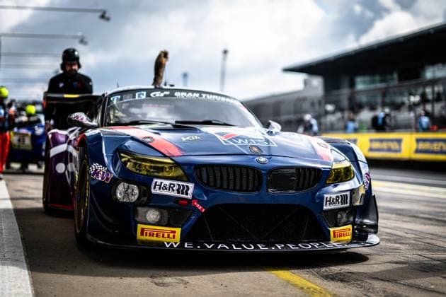 A dark blue BMW Z4 GT3 sponsored by H R, KW and Pirelli is parked in the pit lane of the Nürburgring The vehicle bears the lettering WE VALUE PEOPLE on the front spoiler and the striking WS Racing logo on the hood Team members and the pit area of the 24-hour race can be seen in the background The start number is not clearly legible in the front area BMW Z4 GT3 from WS Racing in the pit lane at the Nürburgring Copyright: xMichaelxGelhardtx