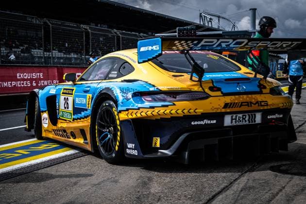 The Mercedes AMG GT3 with the starting number 26 of the PROsport Racing team is parked in the pit lane of the Nürburgring The vehicle Adam Christodoulou Christopher Lulham Marek Böckmann Mikaël Grenier PROsport Racing presents a striking blue and gold pixel pattern design with sponsors such as RAVENOL, Bilstein, DEKRA, Goodyear and H R The pit wall with the PROsport lettering and track marshals can be seen in the background Mercedes AMG GT3 26 from PROsport Racing in the pit lane Copyright: xMichaelxGelhardtx