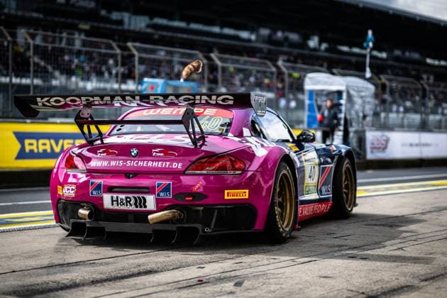 The magenta-colored BMW Z4 GT3 Frank Nikolaus Michael Funke Peter Posavac W I S Racing Team powered by Koopman Racing of the W I S Racing Team powered by Koopman Racing is parked in the pit lane of the Nürburgring The strikingly painted car with the golden rims and the large rear spoiler bears the starting number 40 and shows sponsors such as H R, Pirelli and W I S Safety The filled grandstands and the pit wall with the RAVENOL sponsor banner of the 24h Nürburgring 2026 can be seen in the background BMW Z4 GT3 40 by W I S Racing Team in the pit lane at the Nürburgring Copyright: xMichaelxGelhardtx