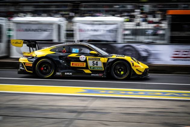 The Porsche 911 GT3 with starting number 54 from Dinamic GT drives through the pit lane at the Nürburgring at high speed The black and yellow car Bastian Buss Michael Christensen Michael Klitgaard Christensen Dinamic GT bears the sponsors RAVENOL, Pirelli and DEKRA prominently on the flank The pit walls and spectator stands can be seen in the background Porsche 911 GT3 54 from Dinamic GT in the pit lane at the Nürburgring Copyright: xMichaelxGelhardtx