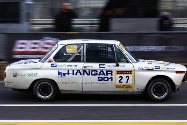 A historic BMW 2002 with starting number 27 passes the pit wall in the pit lane of the Nürburgring The white vehicle bears the inscription Hangar 901 Aircraft Maintenance as the main sponsor as well as the logos of s2 software, RAVENOL and UA Racing The name Bernhard Hagen can be read on the door frame in the cockpit The vehicle takes part in the DHLM German Historic Endurance Championship and shows clear signs of wear from racing Classic BMW 27 with Hangar 901 sponsorship in the pit lane Copyright: xspeedshotx