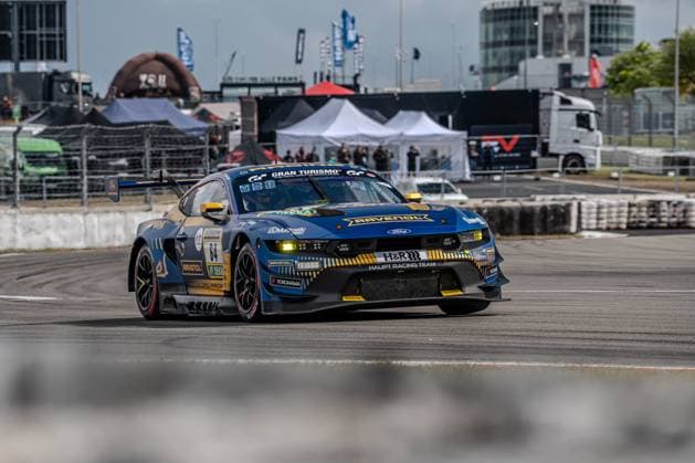 The Ford Mustang GT3 with starting number 64 from HRT Ford Racing drives through a bend on the Nürburgring The dark blue and gold vehicle bears the sponsors RAVENOL, H R and Yokohama and features the Gran Turismo logo on the hood The characteristic buildings of the Nürburgring paddock and hospitality tents can be seen in the background Christopher Mies Dennis Olsen Emil Christian Gjerdrum Finn Wiebelhaus Frederic Vervisch HRT Ford Racing Ford Mustang GT3 64 from HRT Ford Racing at the Nürburgring Copyright: xMichaelxSchäferx