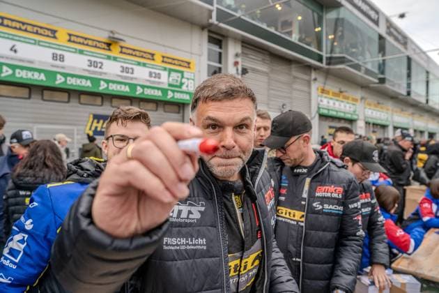 In the Nürburgring paddock, a man in a jacket with the words FRISCH Licht Technische LED Beleuchtung and Giti Competition Tires holds a red pen directly into the camera In the background, other people in team clothing with the emka Motor Oils and SACHS Performance logos can be seen RAVENOL and DEKRA banners as well as start number signs 88, 302, 303, 830 can be seen on the pit garage doors Team representative with pen in the paddock at the Nürburgring Copyright: xMichaelxSchäferx