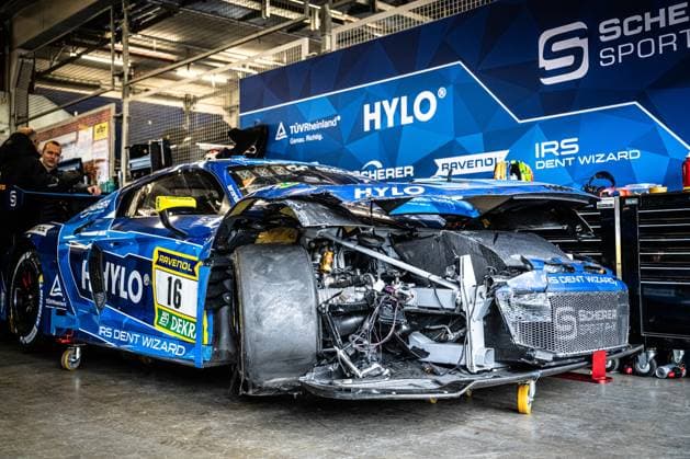 The Alexander Sims Ben Green Christopher Haase Scherer Sport PHX from Scherer Sport PHX is in the garage at the Nürburgring after a serious accident The front of the blue Audi R8 GT3 is completely demolished - body parts are missing, the front area is open, Cables and structural parts are exposed The underbody and the aerodynamic components are heavily deformed The team wall with the sponsors HYLO, RAVENOL, TÜV Rheinland and IRS Dent Wizard is visible in the background A team member observes the vehicle in the background Heavily damaged Audi R8 GT3 16 from Scherer Sport PHX in the pits Copyright: xMichaelxGelhardtx