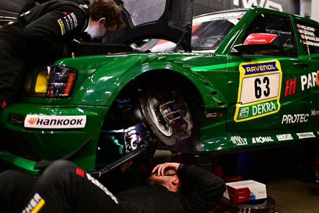 The green racing car Adam Adelson James Green Lance David Arnold Renger Van der Zande HWA Engineering Speed from HWA Engineering Speed stands on lifting jacks in the pit garage at the Nürburgring Two mechanics work intently on the exposed brake disc of the front axle - one kneels underneath, The brake system is completely dismantled and clearly visible Sponsors such as Hankook, RAVENOL, DEKRA and PROTEC can be seen on the bodywork The driver names Holstad, Ludwig, Spengler and Gebhardt are listed on the roof of the vehicle HWA Engineering Speed 63 - Mechanics working on the brakes in the pits Copyright: xMichaelxGelhardtx