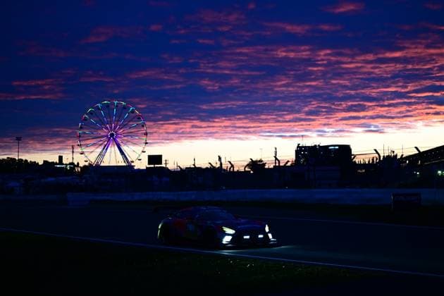 A GT3 racing car crosses the Nürburgring track at sunset The cars headlights shine in the dusk, while a colorfully illuminated Ferris wheel and the silhouette of the paddock infrastructure can be seen on the horizon The dramatically colored sky with orange and purple clouds gives the scene a special atmosphere GT3 car at sunset at the Nürburgring with Ferris wheel Copyright: xMichaelxGelhardtx