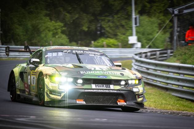 The Ford Mustang GT3 Arjun Maini Fabio Scherer Frank Stippler HRT Ford Racing by HRT Ford Racing drives through a corner on the Nordschleife at the Nürburgring with its headlights switched on The vehicle wears a striking camouflage livery in green and brown tones with the Bundeswehr sponsorship Visible sponsors include H R, Yokohama, RAVENOL and Sparco The typical crash barriers and the lush green of the Eifel can be seen in the background Ford Mustang GT3 67 from HRT Ford Racing on the Nordschleife Copyright: xMichaelxGelhardtx