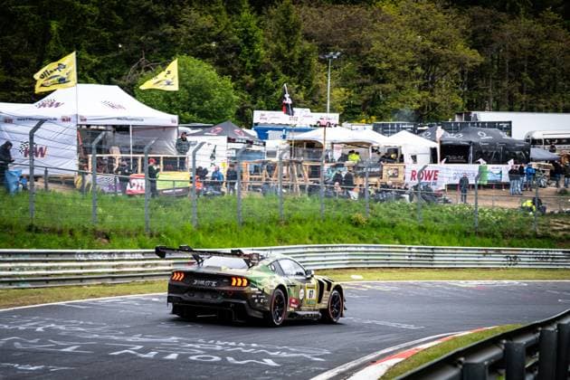The Ford Mustang GT3 from HRT Ford Racing negotiates a corner on the Nordschleife at the Nürburgring The car Arjun Maini Fabio Scherer Frank Stippler HRT Ford Racing wears a striking camouflage livery in olive and black tones and can be seen from behind, with the large rear wing and illuminated brake lights clearly visible In the background are numerous spectators, team tents and advertising banners - including KW and ROWE Motor Oil Ford Mustang GT3 67 from HRT Ford Racing on the Nordschleife Copyright: xMichaelxGelhardtx