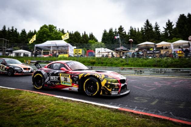 The BMW Z4 GT3 Christian Scherer Henry Walkenhorst Julian Reeh Ralf Schall Saugmotoren Motorsport 3M from Saugmotoren Motorsport 3M takes a corner on the Nordschleife of the Nürburgring The car wears a striking red, black and yellow camouflage design with sponsors such as W S T Welding Specialists Team, RAVENOL, 3M and DEKRA Spectators, tents and the typical Eifel landscape with dense coniferous forests can be seen in the background Another vehicle can be seen in the background BMW Z4 GT3 36 from Saugmotoren Motorsport 3M on the Nordschleife Copyright: xMichaelxGelhardtx