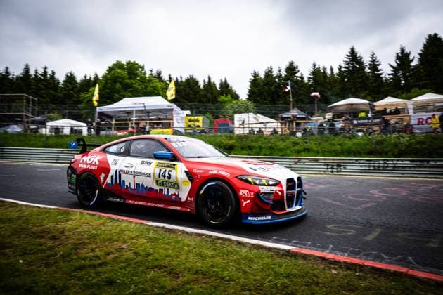 The BMW M4 GT4 Aris Balanian Jeroen Bleekemolen Joshua Bednarski Peter Cate Tom Schütze Riller Schnauck powered by Cerny Motorsport from Riller Schnauck powered by Cerny Motorsport drives through a corner section on the Nordschleife of the Nürburgring The car wears a striking red and white city silhouette design with the sponsors NGK Spark Plugs, RAVENOL and Falken as well as the DEKRA logo In the background, spectator areas and tents can be seen under an overcast sky BMW M4 GT4 145 from Riller Schnauck powered by Cerny Motorsport on the Nordschleife Copyright: xMichaelxGelhardtx