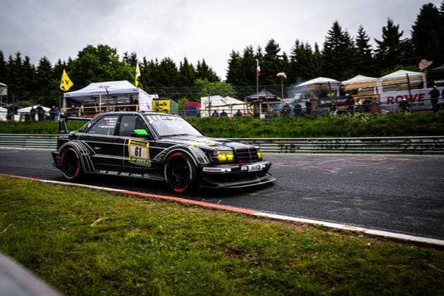 The black-painted Mercedes Benz racing car Bruno Spengler Christian Gebhardt Evald Holstad Peter Ludwig HWA Engineering Speed by HWA Engineering Speed drives through a corner at the Nürburgring The strikingly designed vehicle with yellow headlights and eye-catching aerodynamics package bears the starting number 61 with RAVENOL and DEKRA sponsorship Spectators, tents and the ROWE banner of the paddock can be seen in the background Mercedes Benz 61 by HWA Engineering Speed at the Nürburgring Copyright: xMichaelxGelhardtx