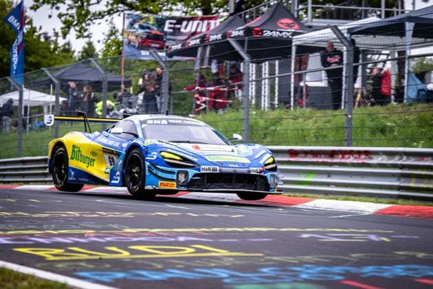 The McLaren GT3 Ben Doerr Marvin Kirchhöfer Timo Glock Timo Scheider Doerr Motorsport from Doerr Motorsport drives through a curve on the Nordschleife of the Nürburgring The vehicle wears the eye-catching blue and yellow Bitburger livery and is equipped with LED headlights Sponsors such as H R and Pirelli are clearly visible In the background, numerous spectators watch the race McLaren GT3 69 from Doerr Motorsport at the Nürburgring Copyright: xMichaelxGelhardtx