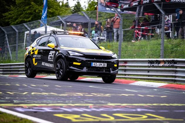 A black and yellow Cupra Race Management vehicle Race Management 3 drives on the Nordschleife of the Nürburgring The vehicle is equipped with an orange blue light bar on the roof and bears the logos of RAVENOL, Löhr Group and the 24h Nürburgring In the background you can see track marshals, fans at the barrier and advertising banners Race Management vehicle on the Nordschleife at the 24h Nürburgring Copyright: xMichaelxGelhardtx