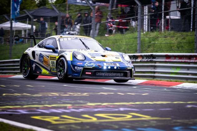 The Porsche 911 GT3 from Clickversicherung de TEAM Kersten Jodexnis Peter Scharmach Richard Jodexnis Richard Sven Jodexnis Robin Chrzanowski Clickversicherung de TEAM takes the red curbs of the Nürburgring in a dynamic cornering maneuver The white and blue car bears sponsors such as RAVENOL, MOTUL and OnlineVersicherung de and is clearly recognizable with the starting number 919 In the background, spectators behind the fences follow the racing action Porsche 911 GT3 919 from Clickversicherung de TEAM on the Nordschleife Copyright: xMichaelxGelhardtx