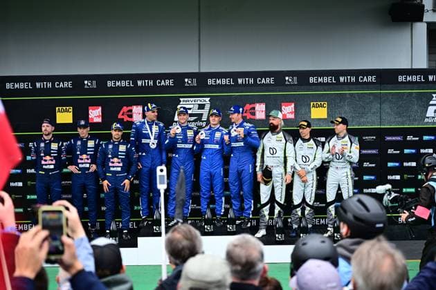 Award ceremony on the podium of the 24h Nürburgring with drivers in blue and white racing suits The teams present their trophies and medals in front of the Bembel with Care banner and sponsor wall Spectators take photos of the winning drivers in the paddock Award ceremony 24h Nürburgring 2026 with drivers and teams on the podium Copyright: xMichaelxGelhardtx