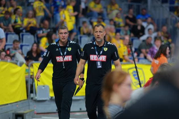 Coach Maik Machulla and co-coach Michel Abt of the Rhein Neckar Löwen run in black team outfits with REWE sponsor logo on the sideline of the SAP Arena The yellow-clad home fans can be seen in the background Maik Machulla and Michel Abt Rhein Neckar Löwen on the sideline Copyright: xspeedshotx