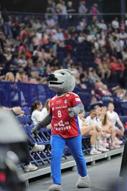 The HSV mascot in red and blue costume waves to the spectators in the Barclays Arena In the background you can see players in blue HSV jerseys and fans in the stands HSV Hamburg mascot looking after players Copyright: xspeedshotx