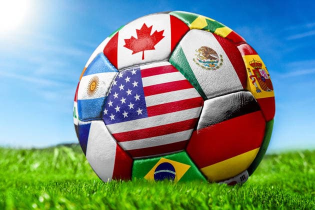 FOTOMONTAGE, Symbolfoto zur Fußball-Weltmeisterschaft 2026 - Fußball mit verschiedenen Nationalflaggen auf Spielfeld Rasen unter blauem Himmel. Zu sehen sind unter anderem die Flaggen der USA, Mexikos, Kanadas, Spaniens, Brasiliens und Argentiniens.