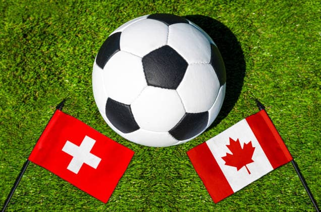 FOTOMONTAGE, Fußball-Weltmeisterschaft 2026: Nationalflaggen von Schweiz und Kanada mit Fußball auf Rasen. Symbolbild zum WM-Gruppenspiel am 24 Juni bei der FIFA Fussball-Weltmeisterschaft 2026