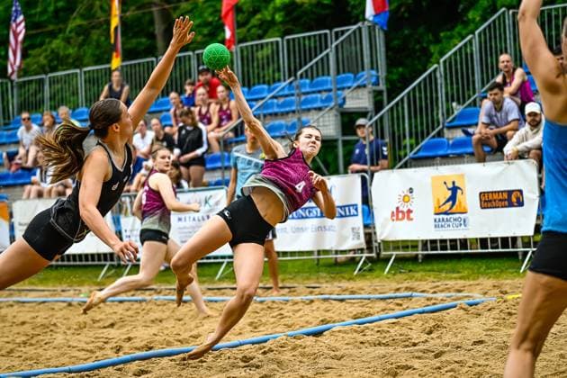 A player in the pink jersey 4 throws at the goal from an extreme angle at the German BeachOpen Karacho Beach Cup, while two opponents in black jerseys try to block the shot In the background, spectators watch the game from the stands Player 4 attempting to throw at the Karacho Beach Cup Copyright: xspeedshotx xDorianfrickx