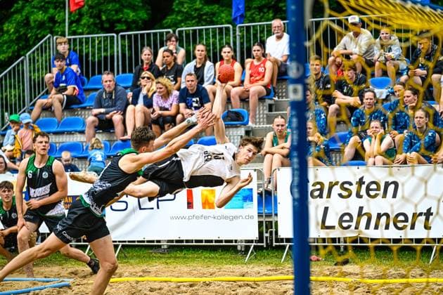 A player in a white jersey 10 throws himself towards the goal in an acrobatic flying movement, while an opponent in a black jersey tries to block the shot The scene takes place on a sand court in front of a packed crowd Spectacular throw attempt in beach handball in front of a full crowd Copyright: xspeedshotx xDorianfrickx
