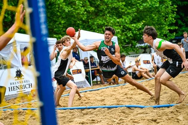 A player in the green and black jersey with the number 10 takes a shot from the back and is blocked by an opponent The scene takes place on an outdoor sand court, with spectators and advertising boards visible in the background Player 10 attempting a throw at a beach handball tournament Copyright: xspeedshotx xDorianfrickx