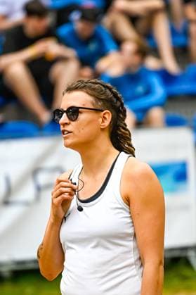 A woman with sunglasses and braided hair stands on the sideline holding a microphone or headset In the background you can see blue grandstand seats with spectators Coach or official on the sideline at the outdoor event Copyright: xspeedshotx xDorianfrickx