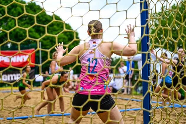 A goalkeeper in a pink Germany jersey with the number 12 stands in the goal with her arms outstretched and watches the game The picture was taken through the goal net, in the background you can see players in green jerseys Goalkeeper 12 Germany holds the goal during beach handball Copyright: xspeedshotx xDorianfrickx