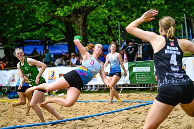 A player with the number 15 throws at the goal while an opponent of Beach Unicorns 4 prepares the defense In the background, a player with the number 12 in a green jersey follows the scene on the sand court Player 15 throwing against Beach Unicorns 4 Copyright: xspeedshotx xDorianfrickx