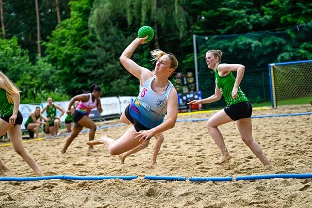 A player with the number 15 in a white jersey jumps into the air during a beach handball game and throws the green ball at the goal Opposing players in green jerseys try to stop the attack Player 15 throwing the ball in beach handball Copyright: xspeedshotx xDorianfrickx