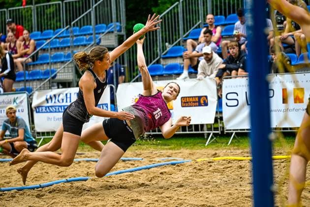 Player 15 in the pink jersey throws at the goal in a spectacular flying movement at the German Beach Open beach handball tournament, while an opponent in the black jersey tries to block the ball The green handball is clearly visible in the picture Spectacular throwing action 15 at the German Beach Open Copyright: xspeedshotx xDorianfrickx