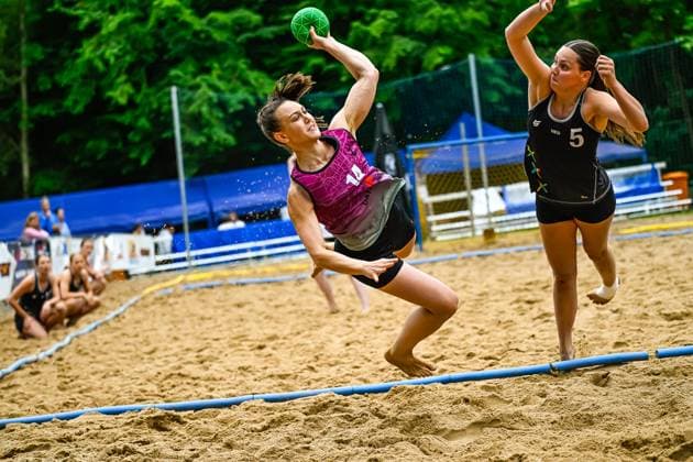 A player with the number 14 jumps dynamically and throws the green ball towards the goal, while an opponent from the Netherlands 5 tries to block the throw The game takes place on an outdoor sand court Player 14 attempting to throw in a beach handball game Copyright: xspeedshotx xDorianfrickx