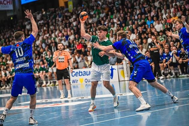 Elias Newel 40 Frisch Auf Göppingen draws up to shoot while Ivan Snajder 10 TVB Stuttgart blocks on the left and another TVB defender tries to disrupt the shot In the background, numerous spectators watch the scene in a packed arena Elias Newel 40 Frisch Auf Göppingen taking a shot against TVB Stuttgart Copyright: xspeedshotx xDorianfrickx