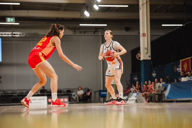 A player in a white jersey is dribbling the ball and looking for a teammate to pass to, while an opposing player in a red jersey with the number 12 is blocking her path. The scene takes place at Kistamässan in Stockholm. A player with the ball in a one-on-one situation during a basketball game in Stockholm Copyright: xJoelxKruehlerx