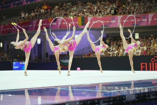 The Spanish Ensemble group performs a synchronized lift routine with hoops and clubs on the competition floor of the Festhalle in Frankfurt. The scoreboard displays the team name Spain, and in the background, a packed audience watches the Spanish Ensemble group’s performance with hoops and clubs at the World Championships in Frankfurt. Copyright: xspeedshotx