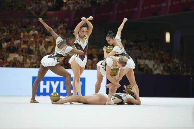 A group of five gymnasts performs a synchronized group routine with golden balls on the white competition mat at the Festhalle in Frankfurt. Several athletes raise their fists in the air, while one gymnast lies on the floor and another bends down deeply. Group routine with golden balls at the Rhythmic Gymnastics World Championships in Frankfurt Copyright: xspeedshotx