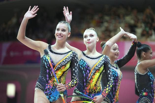 Three gymnasts from a group team smile and wave to the audience in Frankfurt’s Festhalle while holding clubs and hoops. In the background, a fourth athlete can be seen with a Portuguese flag on her leotard. 2026 Rhythmic Gymnastics World Championships – Group team waves to the audience Copyright: xspeedshotx
