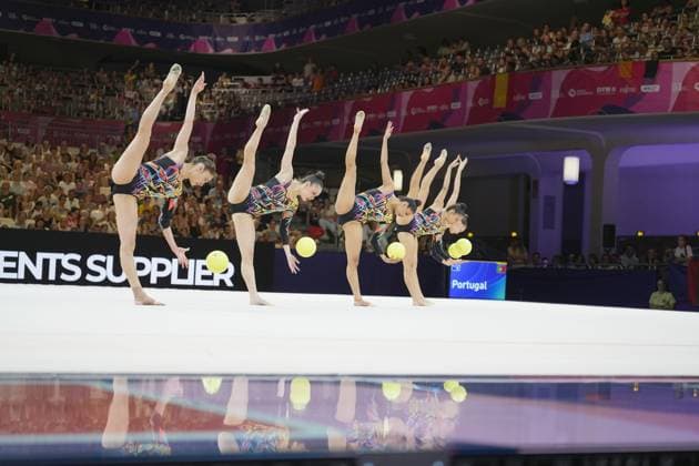 The Portuguese team performs a synchronized balancing routine with yellow balls, with all five gymnasts raising their straight legs vertically upward. The scene is reflected in the gleaming floor of the Festhalle Frankfurt. Portugal during the group routine with balls – Rhythmic Gymnastics World Championships, Frankfurt Copyright: xspeedshotx