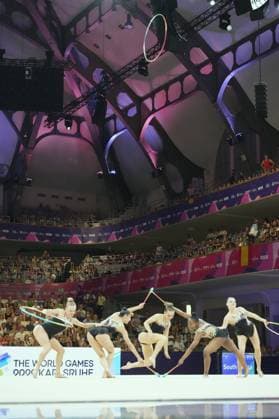 A group of five gymnasts performs a synchronized group routine with hoops and clubs at the Festhalle in Frankfurt. A hoop is thrown high into the air, while the athletes perform in coordinated positions on the competition floor. Group routine with hoops and clubs at the Rhythmic Gymnastics World Championships in Frankfurt Copyright: xspeedshotx