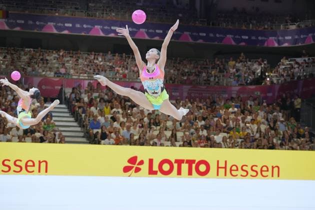 Two gymnasts perform a synchronized jump combination with pink balls in Frankfurt’s packed Festhalle. The gymnast in the front performs a straddle jump while the ball is thrown into the air. Group routine with a ball at the Rhythmic Gymnastics World Championships in Frankfurt Copyright: xspeedshotx
